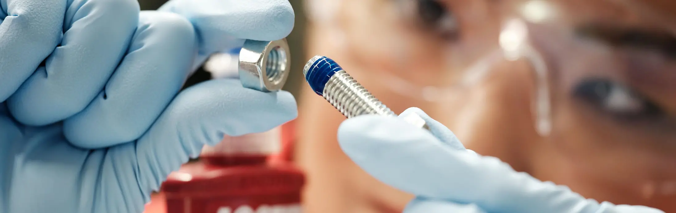 En el fondo, el rostro de una mujer. Al frente, sus manos con guantes azules sosteniendo un tornillo con Threadlocker azul.