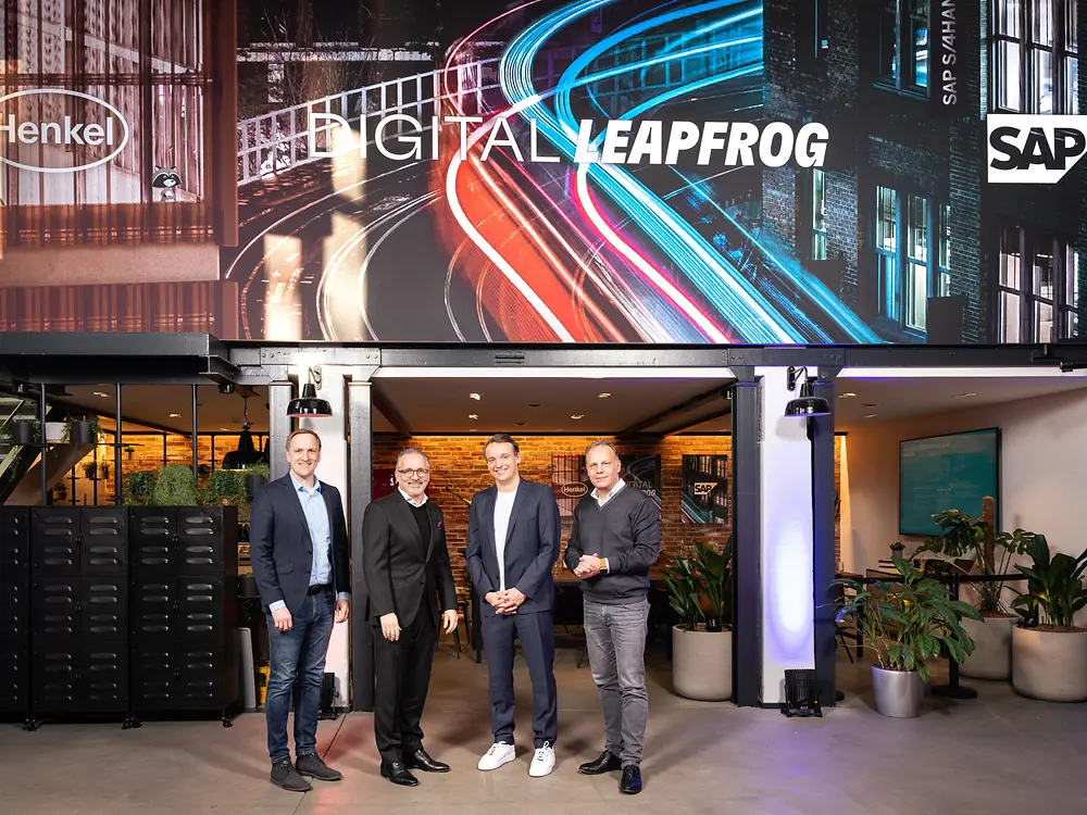 Four men standing in front a branded wall showing the Henkel and SAP logo. 